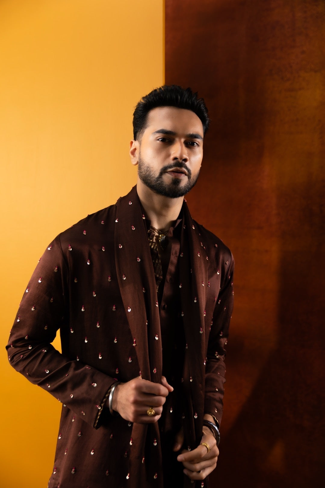 A man wearing a dark brown embroidered kurta with small metallic motifs, posed against a split yellow and dark brown studio background.