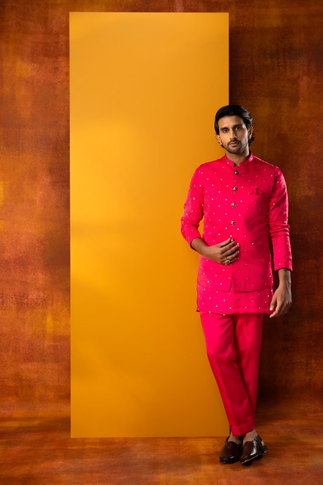 A man modeling a bright pink traditional Indian outfit with small white embroidered details, standing against a textured brown backdrop with a large yellow rectangular panel behind him.