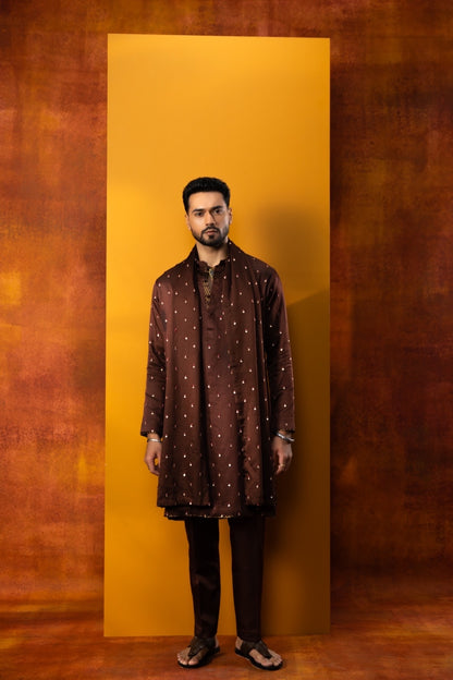 A man stands facing forward in a studio setting, wearing a dark brown long kurta with small white dot patterns, dark trousers, and sandals. He is positioned in front of a tall mustard-yellow rectangular backdrop against a textured brown and orange background.