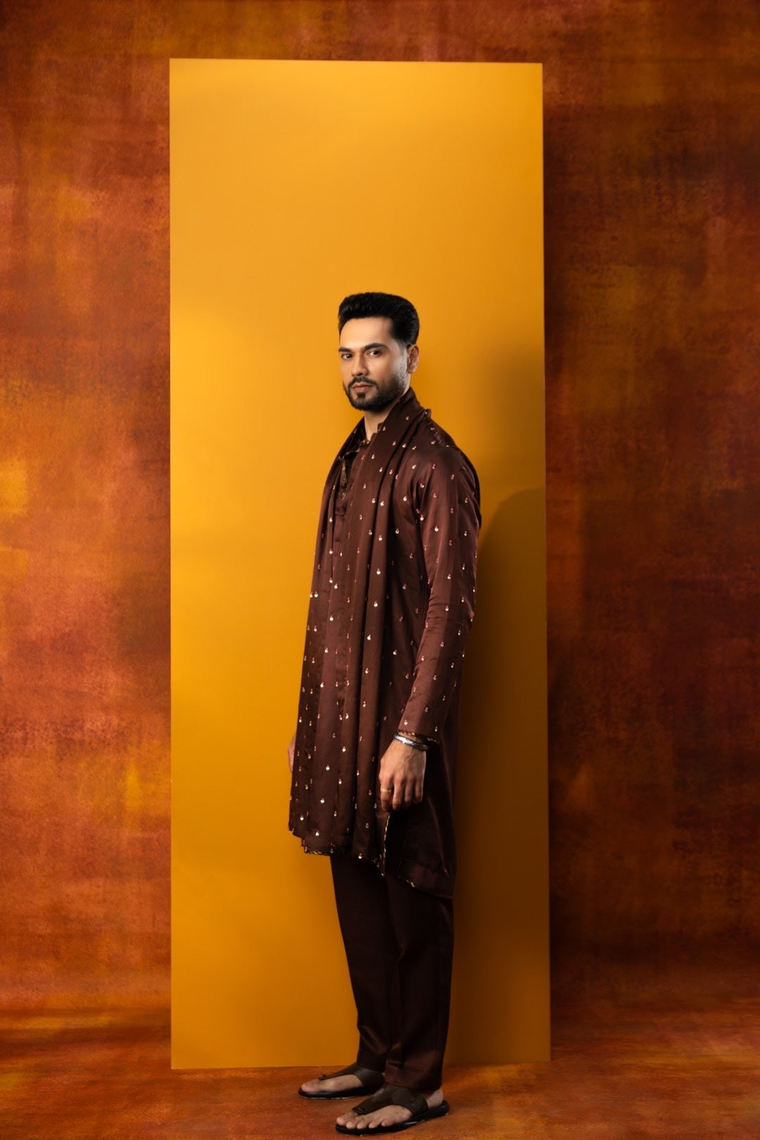 A man wearing a dark brown patterned kurta and dark trousers stands in profile against a large mustard yellow rectangular backdrop, with a textured brown studio background.