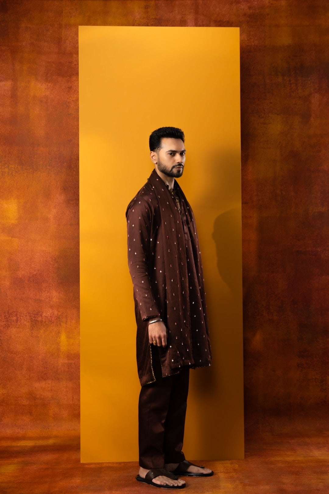 A man wearing a dark brown embroidered kurta and matching trousers stands in profile against a large mustard yellow rectangular backdrop, with a textured brown studio background.