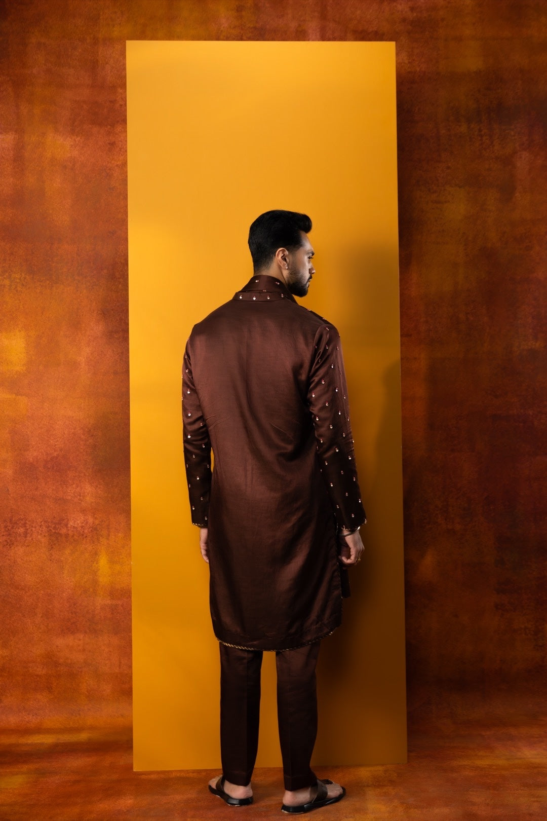 A man wearing a dark brown embroidered kurta and matching trousers, standing with his back to the camera in front of a vertical yellow backdrop against a textured brown background.