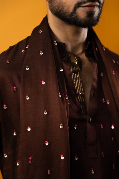 A close-up of a man wearing a dark brown embroidered sherwani jacket with small metallic embellishments, paired with a patterned tie, against a solid orange background.