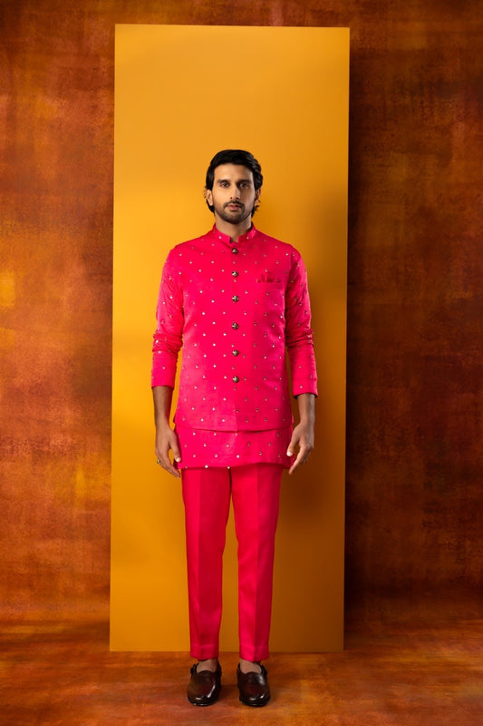 A man modeling a bright pink traditional Indian outfit consisting of a patterned kurta and matching trousers, standing in front of a vertical yellow backdrop against a textured brown background.