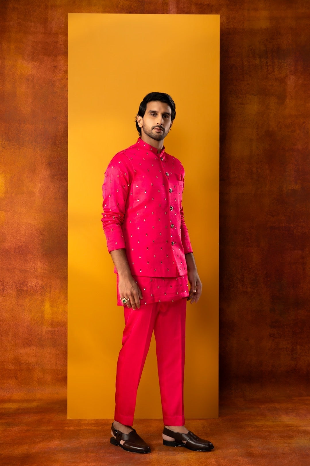 A man modeling a bright pink traditional Indian suit with silver embroidery, consisting of a band-collar jacket, matching trousers, and black loafers, standing in front of a yellow rectangular backdrop against a textured brown background.