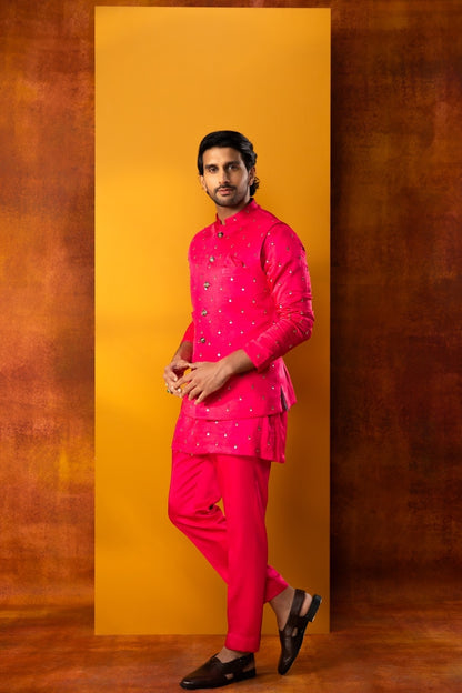 A man modeling a bright pink traditional Indian outfit with small white embroidered details, consisting of a long-sleeve kurta and matching trousers, posed against a warm yellow and brown textured studio backdrop.
