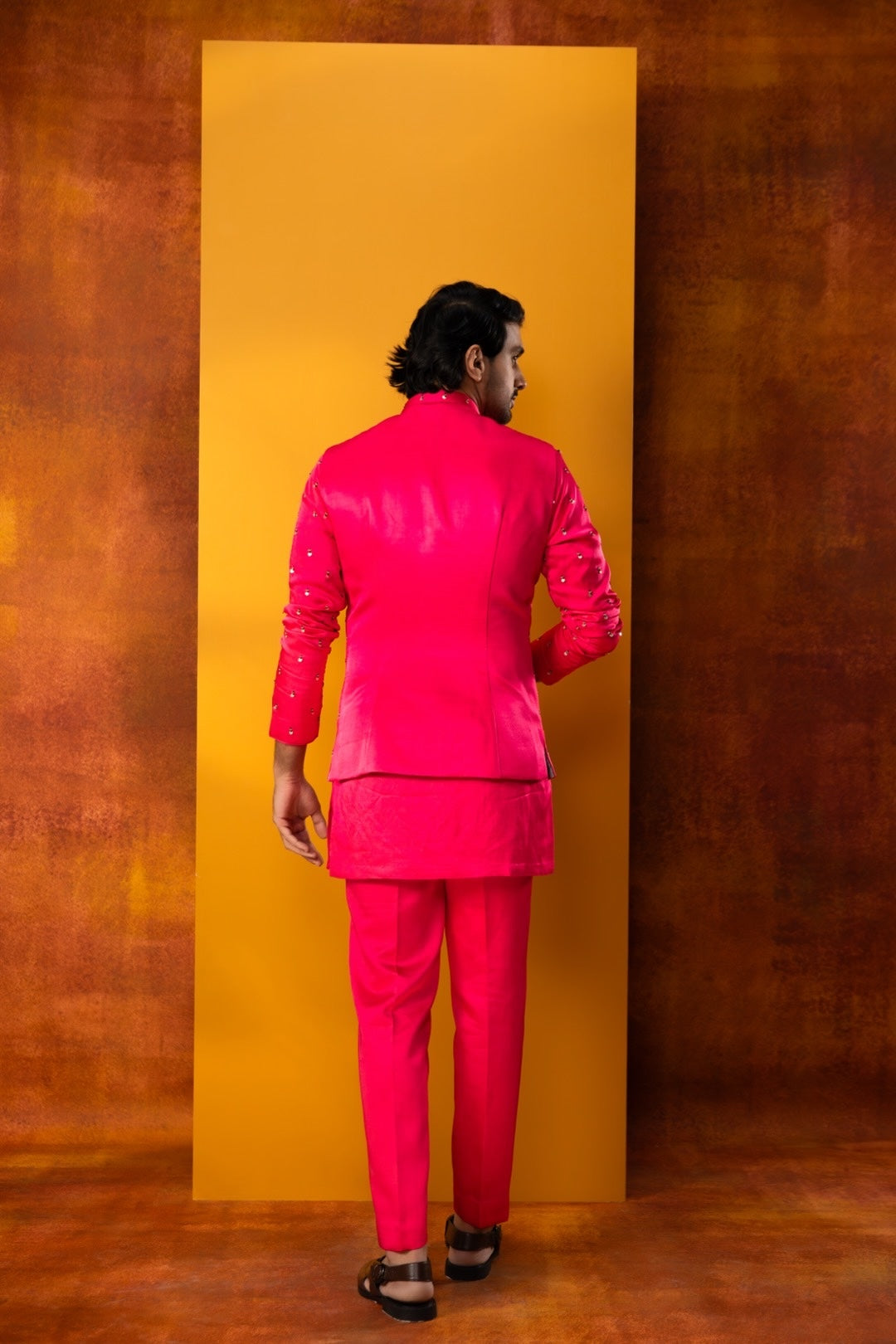 A man wearing a bright pink traditional Indian suit with a matching jacket and trousers, standing with his back to the camera in front of a large yellow rectangular backdrop against a textured brown background.