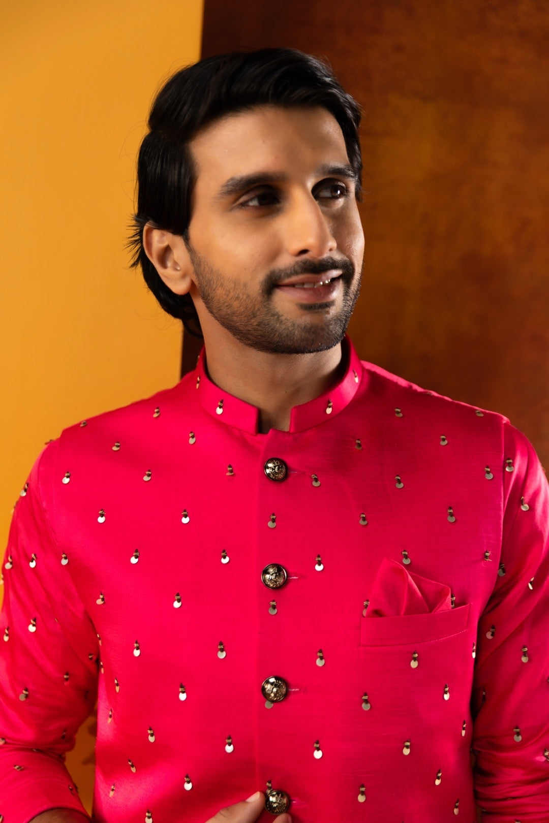 A man wearing a bright pinkish-red sherwani jacket with small metallic embellishments, set against a warm orange and brown background.